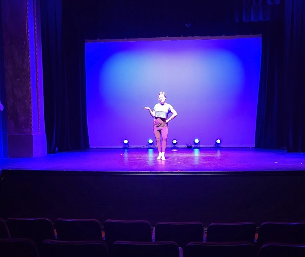person standing on a stage in blue and purple lighting with empty chairs in the foreground