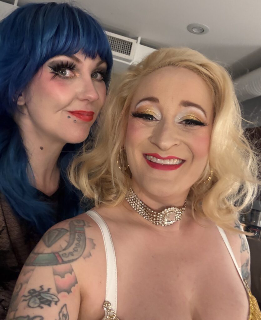 two showgirls backstage, one with blue hair and one blonde